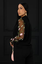 Woman wearing a black outfit with floral embroidery on sheer sleeves against a plain background