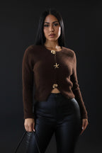 Woman wearing a brown cardigan with decorative buttons and black pants against a plain background